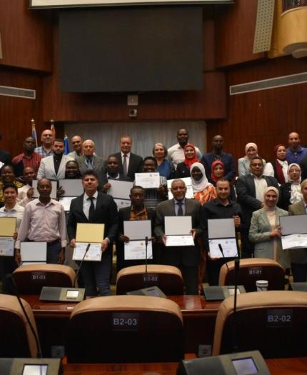 A group of people in formal attire stand in rows holding certificates inside a conference room with wooden walls and numbered seats.