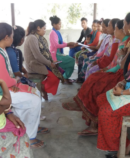 A group of people sit in a circle outdoors, some holding notebooks, engaged in discussion. A child is seated on one person's lap.