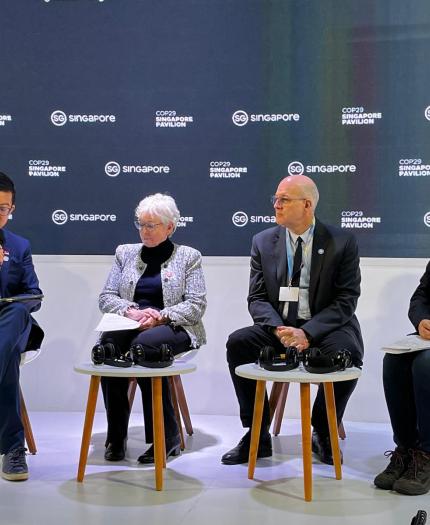 Four individuals are seated on stage, engaging in a panel discussion. Behind them is a backdrop with logos and text. Microphones and headphones are placed on the small tables in front of them.