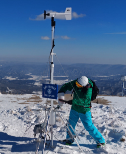 Person in winter gear adjusts a weather station on a snowy mountain summit under a clear blue sky.