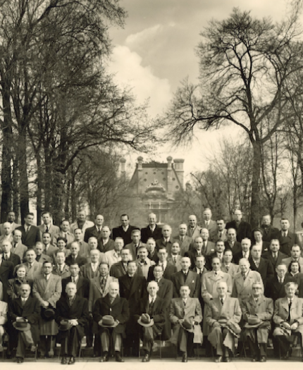 A large group of men in suits and coats pose for a formal photo outdoors, with trees lining a path and a building visible in the background.