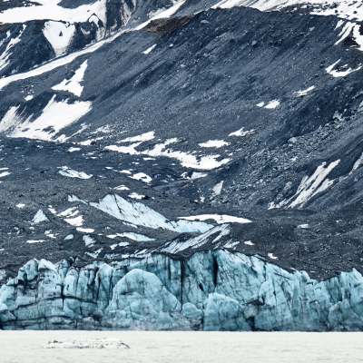 A glacier with blue ice formations is surrounded by rocky, snow-dusted mountains, and a body of water is visible in the foreground.