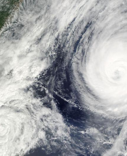 Satellite image of two large cyclones in the ocean near a landmass, with swirling cloud patterns visible.