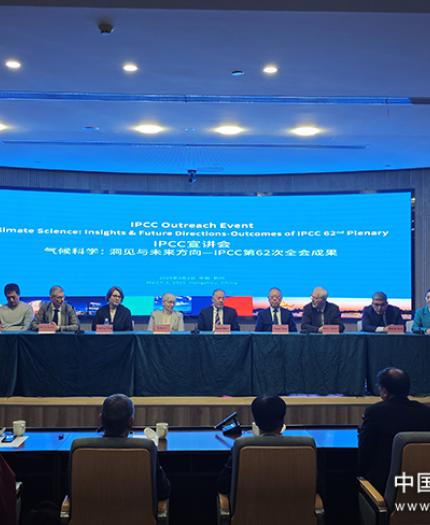 A panel of nine people seated at a table during an IPCC outreach event, with a large blue presentation screen behind them. Audience members are visible in the foreground.
