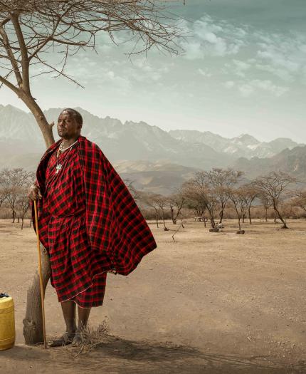 A person in traditional red attire leans against a tree in a dry landscape, with mountains in the background and a yellow container nearby.