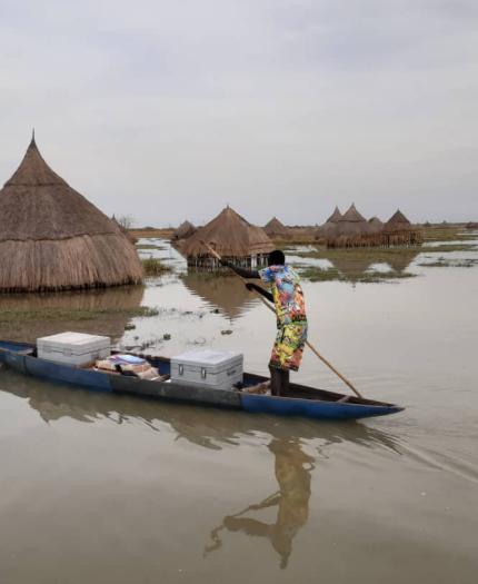 A person paddles a small canoe on a floodplain, passing traditional thatched huts partially submerged in water.