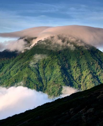 A green mountain peak is partially covered by a cap of clouds, with additional clouds surrounding its base under a clear blue sky.
