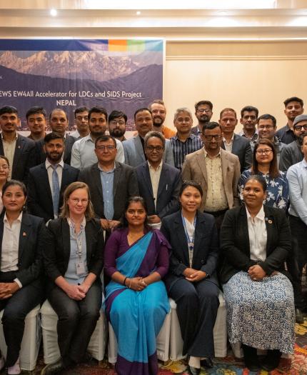 A group of men and women in business attire pose for a photo in a conference room with a banner reading "CREWS EW4All Accelerator for LDCs and SIDS Project Nepal" in the background.
