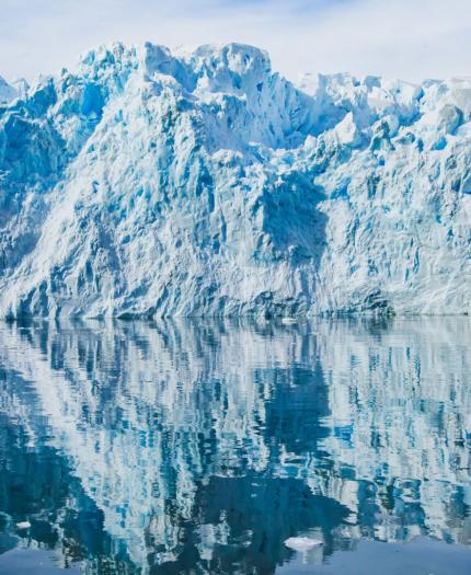 A glacier with jagged blue and white ice is reflected in calm, clear water under a partly cloudy sky.