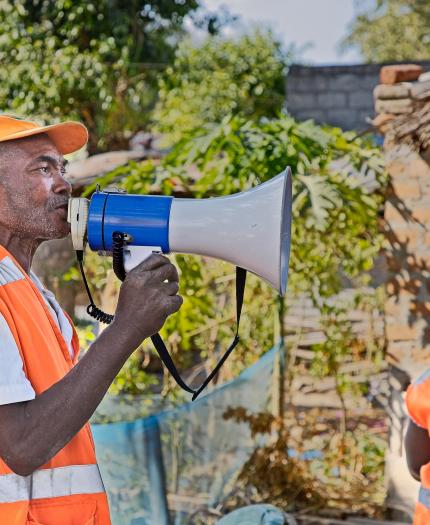 A man in an orange vest and cap speaks into a megaphone outdoors while another person in similar attire stands nearby.