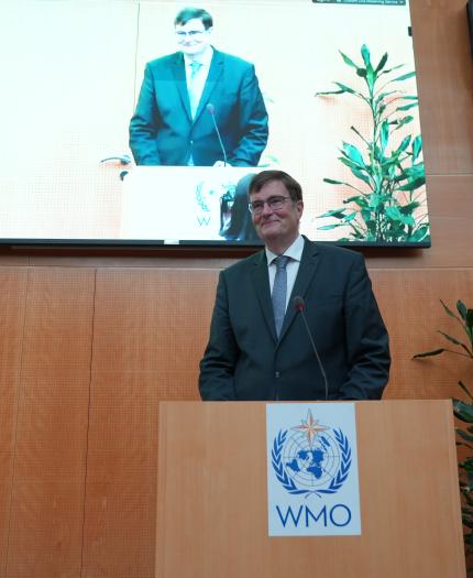A man in a suit stands at a podium with the WMO logo, speaking in front of a screen displaying his image in a wood-paneled room with plants.