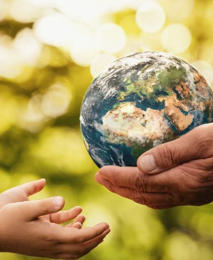 Two adult hands hold a small globe while two child hands reach out towards it, set against a blurred, green outdoor background.
