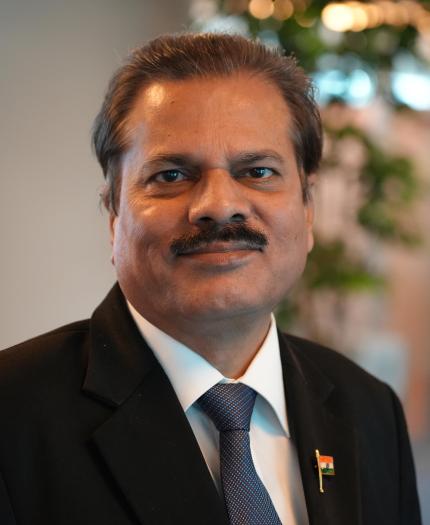 A middle-aged man in a suit and tie with a small Indian flag pin, standing indoors with blurred office background.