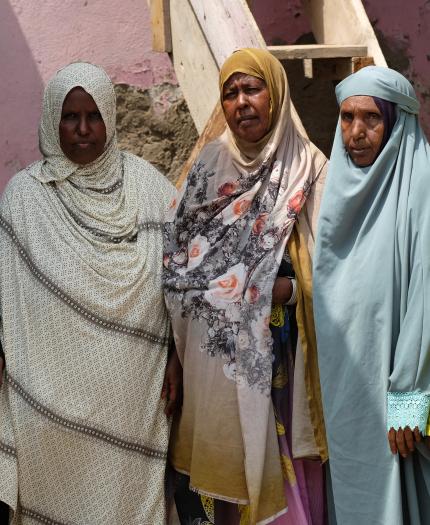 Five women in colorful hijabs stand side by side in front of a pink wall and wooden stairs, looking at the camera.