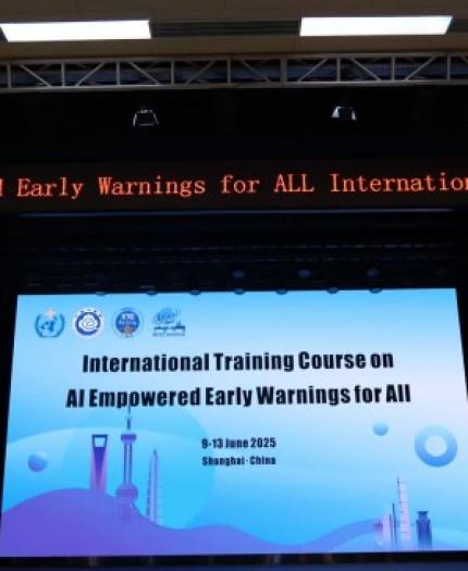 A conference stage displays a banner and digital sign for the "International Training Course on AI Empowered Early Warnings for All," held in Shanghai, China, June 2025.