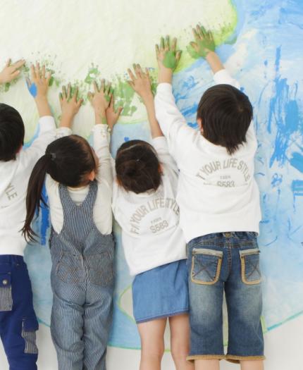 Four children stand facing a wall, pressing their painted hands onto a large drawing of the Earth.