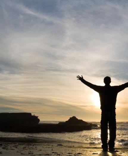 A person stands on a rocky beach with arms outstretched, facing the ocean at sunset under a partly cloudy sky.