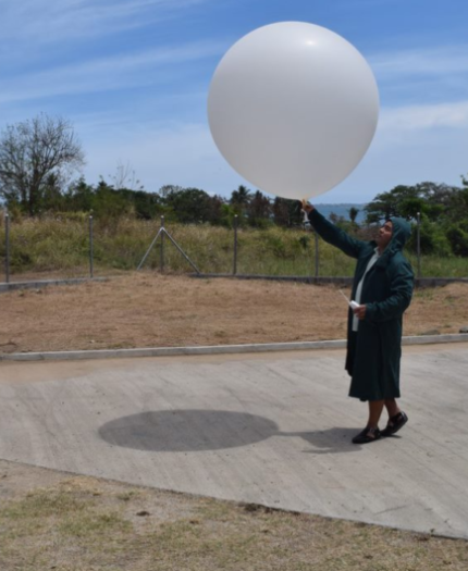 A person in a coat and headscarf stands on a paved area holding a large white weather balloon outdoors under a blue sky.