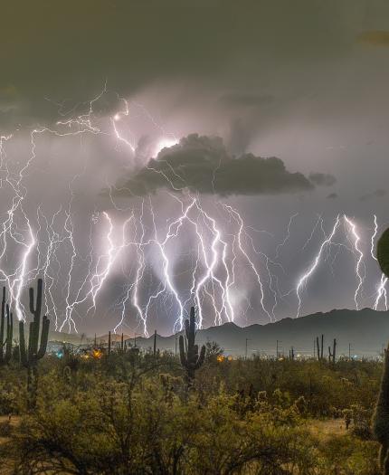 Multiple lightning bolts strike over a desert landscape with cacti and distant mountains at night, illuminating the cloudy sky.