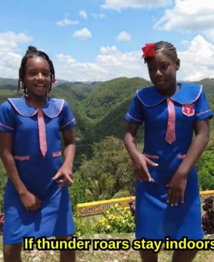 Two girls in blue school uniforms stand outdoors on a hilltop with green hills in the background. Subtitle reads, "If thunder roars stay indoors," in yellow text.