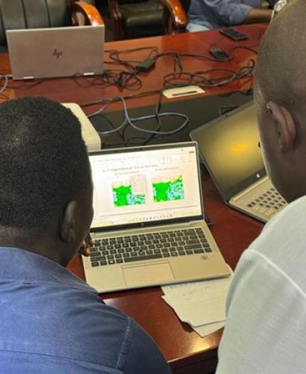 Two people look at weather or map data displayed on a laptop screen in an office meeting setting, with laptops and papers on the table.