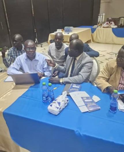 A group of people sit around a table with laptops, notebooks, and bottled water, engaged in discussion at an indoor event or meeting.