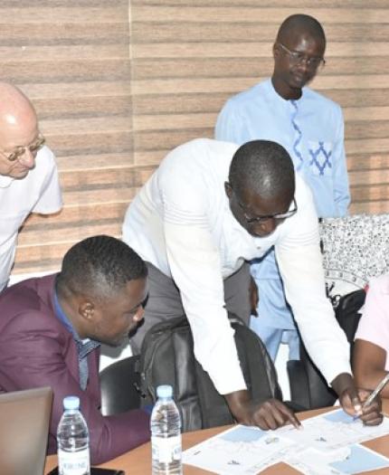 Five people are gathered around a table with papers, a laptop, and water bottles, engaged in a discussion and reviewing documents in an office setting.