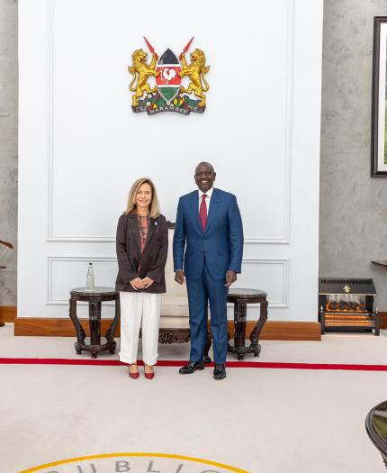 Two people stand side by side in a formal room with framed photographs, decorative furniture, and a national emblem on the wall behind them.