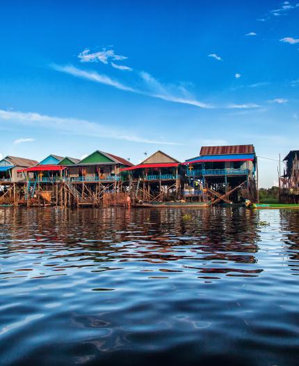 Colorful wooden houses on stilts line the edge of a calm body of water under a blue sky with scattered clouds.