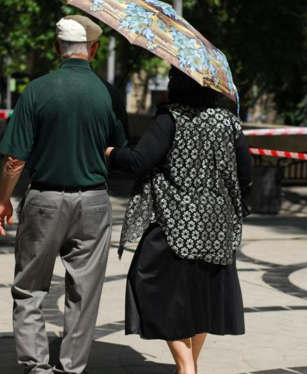 An older couple walks down a sunny, tree-lined sidewalk. The woman holds a large umbrella, and they are walking arm in arm away from the camera.