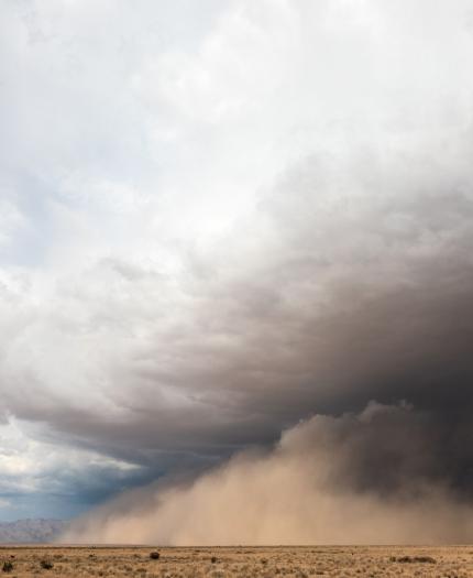 A large dust storm approaches across a dry, flat desert landscape under a partly cloudy sky.