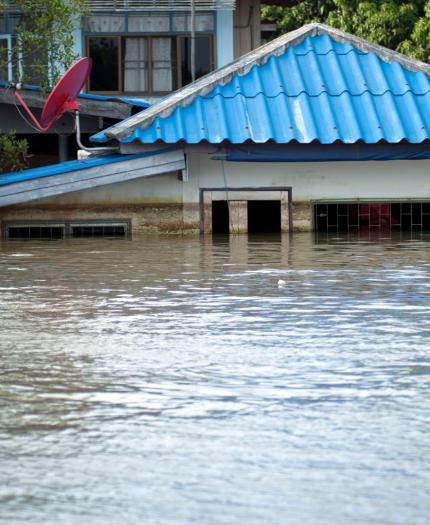 A house with a blue roof is partially submerged in floodwater, with only the roof and upper windows visible above the waterline.