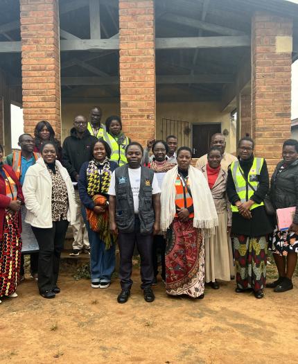 A group of adults stand together outside a brick building, some wearing reflective vests and others in patterned clothing, posing for a group photo.