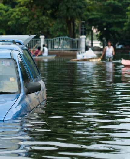 A blue car is partially submerged in floodwater, while people use boats to navigate a flooded street lined with trees in the background.