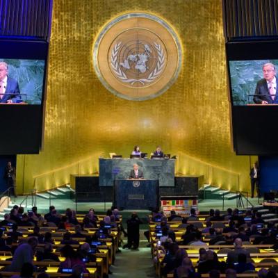 A speaker addresses delegates at the United Nations General Assembly hall, with two screens displaying his image and the UN emblem visible in the background.