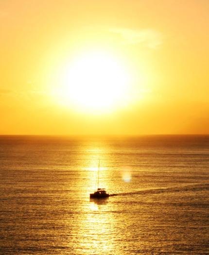 A small boat sails on calm water at sunset, with the sun low on the horizon and golden light reflecting on the sea.