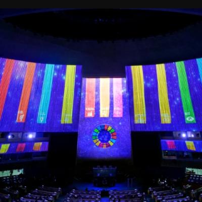 The interior of a large auditorium displays multicolored vertical light panels and a circular emblem on the wall above seated attendees.