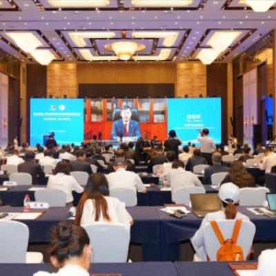 A large conference room filled with attendees facing a stage and screens displaying a speaker’s image during a formal event.