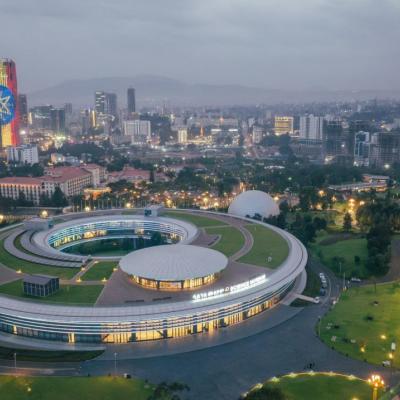 An aerial view of a modern, circular building with a central courtyard, surrounded by greenery and city lights, with a city skyline and tall buildings in the background at dusk.