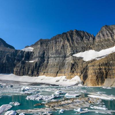 Rocky cliffs rise above a partially frozen turquoise lake with floating ice and patches of snow under a clear blue sky.