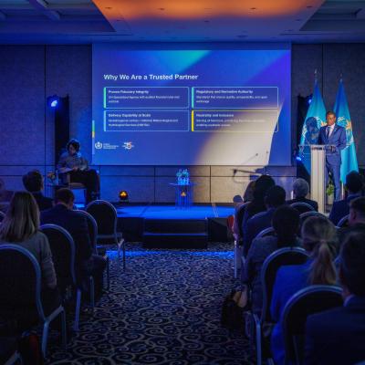 A speaker presents information on a screen titled "Why We Are a Trusted Partner" to an audience seated in a conference room with blue lighting.