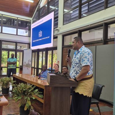 A man speaks at a podium during an indoor event, with attendees seated at tables and banners displaying "Solomon Islands Meteorological Service" visible in the background.