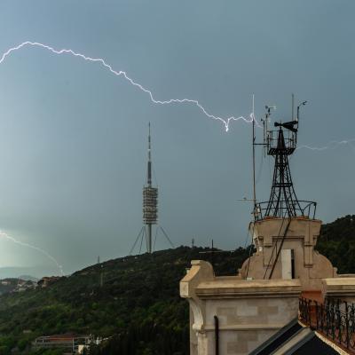A bolt of lightning strikes near a tall communications tower and an observation building on a hill, with dark clouds overhead and green landscape below.