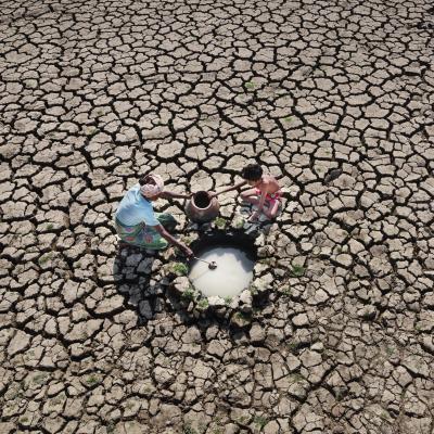 Three people collect water from a small well in the middle of a cracked, dry landscape, indicating drought conditions.
