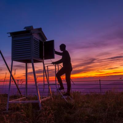 A person checks equipment inside a weather station at sunset, standing on a small ladder beside a fenced area and tall antenna structure.