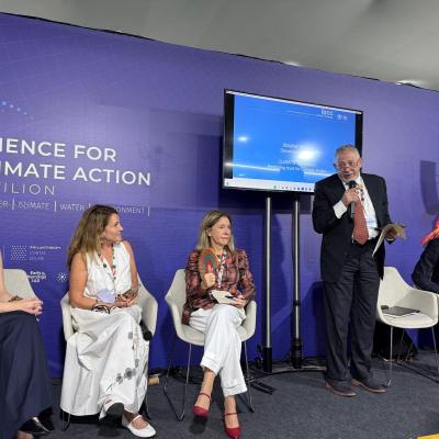 Five panelists sit and stand on a stage at the Science for Climate Action Pavilion, with a man speaking into a microphone and a presentation screen behind them.