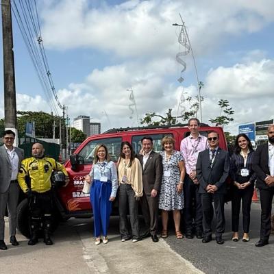 A group of people in business attire stand in a line outdoors in front of a red vehicle, with trees and a partly cloudy sky in the background.