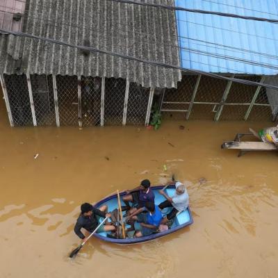 Three people row a small boat through floodwaters in front of submerged buildings, while a man sits on a table with bags nearby.