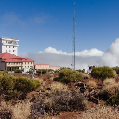 A research observatory building with a red roof, surrounded by dry shrubs and antennas, under a blue sky with scattered clouds.