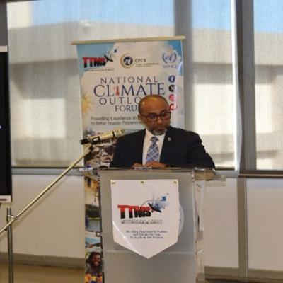 A man in a suit stands at a podium speaking during the National Climate Outlook Forum, with a presentation screen and banner in the background.